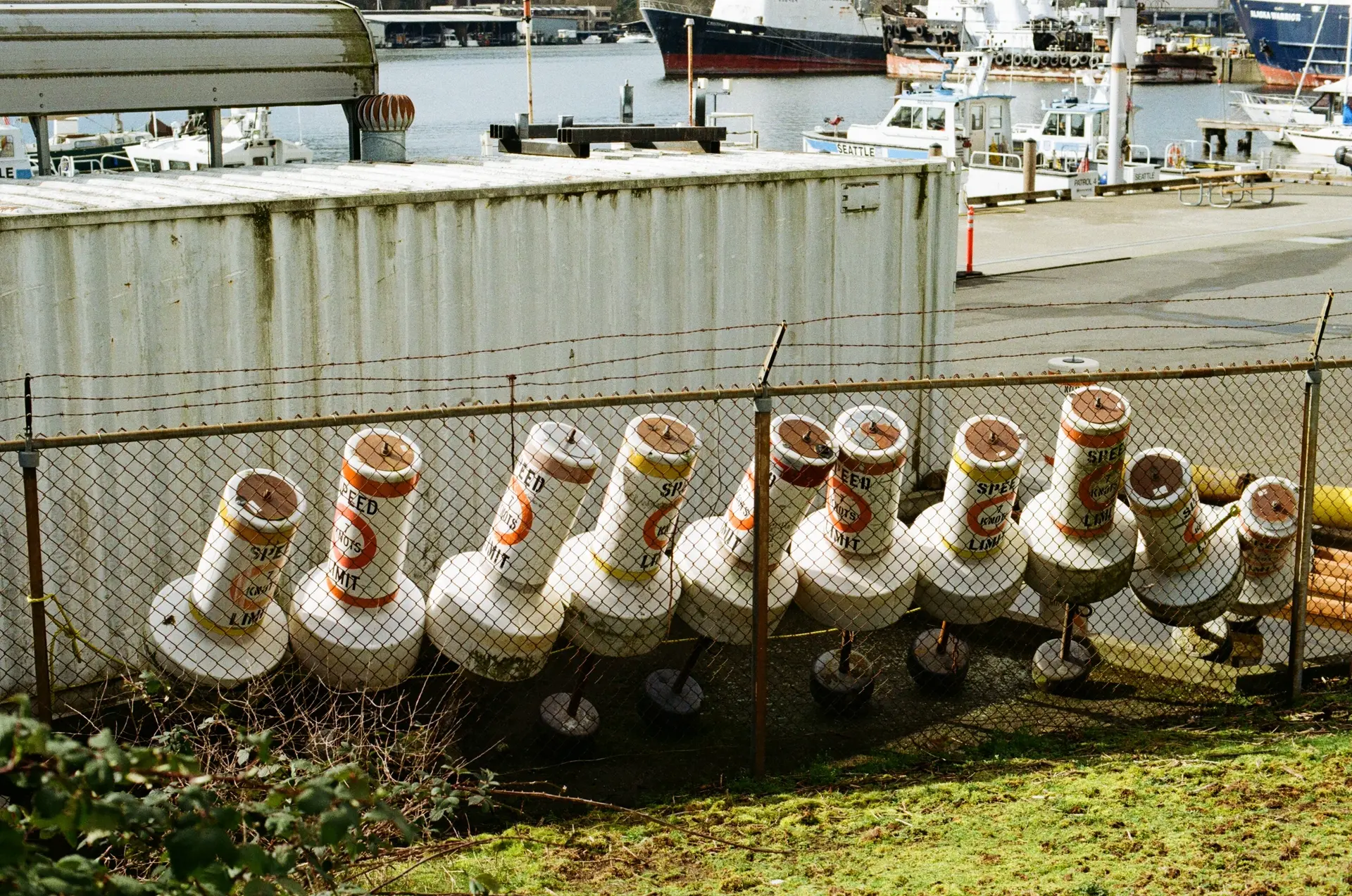 Photograph of speed limit bouys resting in various angles between a white shipping container and a chain link, barbed wired fence. They are in police territory -- in the distance, Seattle PD boats are docked and visible.
